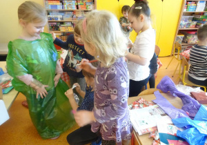 Jedna dziewczynka ma założony duży zielony worek plastikowy, który okrywa jej ciało, do której trzy dziewczynki doklejają elementy dekoracyjne.